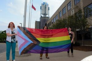 Three women hold a rainbow coloured flag in front of a college building
