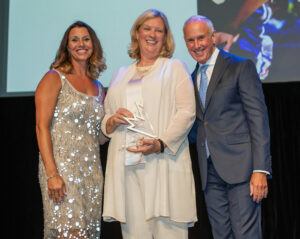 two women and a man dressed in formal wear smile on stage with a large digital screen in the background
