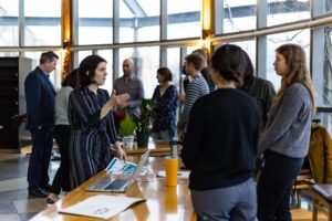 A woman with dark, short hair in a striped dress standing across a table from students engaged in conversation