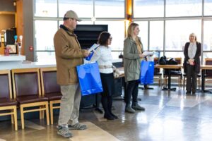 From left to right: A man in a brown coat and hat, a woman with short dark hair and a white shirt and a woman with a green coat and blonde hair. Man on left and woman on right both holding large blue bags.