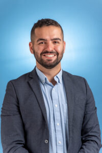 A headshot of a man in a light blue button up and grey blazer, smiling at camera with a blue background.