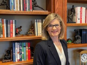 Photo of a woman with short hair wearing glasses, standing in front of a bookshelf smiling at camera