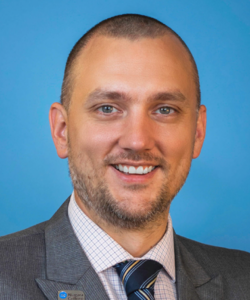 A headshot of a man wearing a suit and tie, smiling and looking at camera with a blue background.