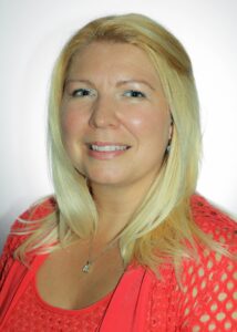 A woman with short blonde hair wearing an orange shirt, smiling at camera in front of white background.