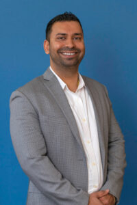 A headshot of a man in a white button up shirt and grey blazer smiling and looking at camera with a blue background.