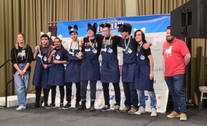 A group of students wearing cat ears and blue aprons, standing in a huddle and smiling in front of a banner wearing medals and holding a gold trophy.