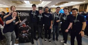 A group of standing standing in a huddle smiling and looking at camera, wearing shirts that read Mosu and medals around their neck and holding a gold trophy.