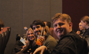 Three people sitting in a row in seats of a dimmed theatre smiling at the camera.