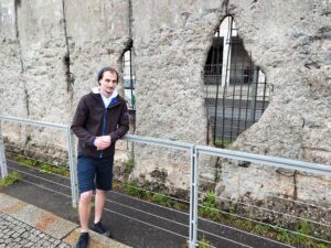 A man leaning on a railing in front of a stone wall, smiling at camera.