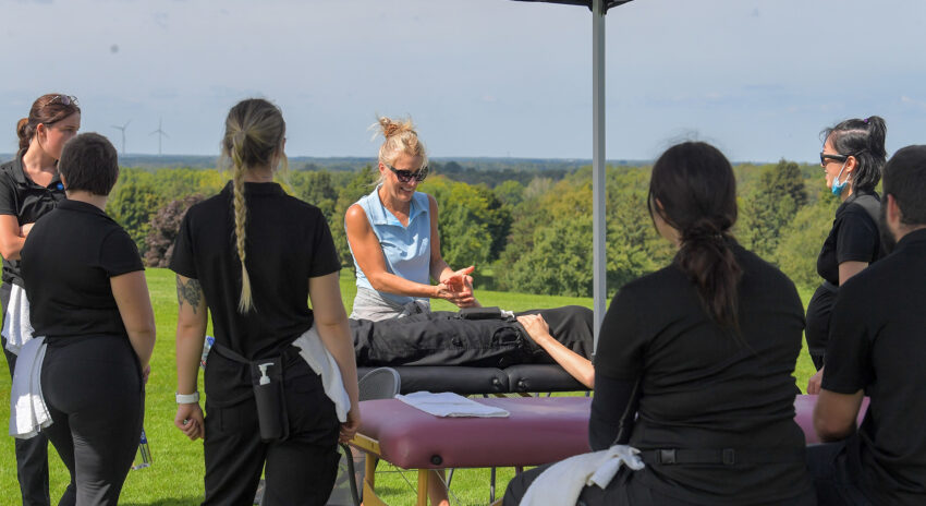 A women standing in front of a table on a golf course, performing a demonstration in front of a group of people.