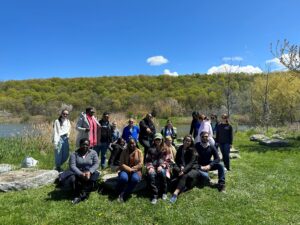 A group of people standing and sitting outdoors on green grass in front of a hill in the background.