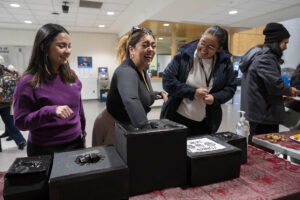 Three women laughing over activitiy taking place on table. Women in the middle has her hand placed in a black box on table.