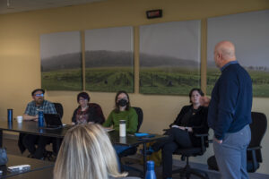 A person in a long sleeve blue shirt facing a panel of four people sitting at a desk in front of a yellow-painted wall and four canvas photos.