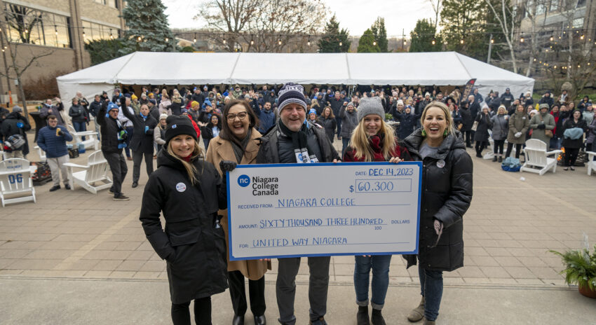 A group of five people standing outside wearing parkas and hats, holding a big cheque.