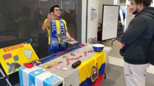 A man standing in front of a table wearing a yellow shirt and blue scarf. Standing beside yellow poster that reads Ecuador.