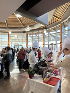People gathered in indoor space with lots of windows. At the forefront is people standing in chef hats and aprons around table with food.