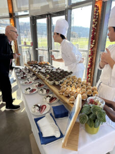 Two people wearing chef hats and aprons standing behind table with several desserts and speaking to a person overlooking table.