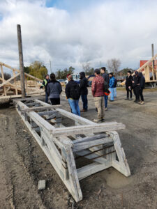 A wooden fixture at forefront with a group of people standing on outdoor construction site in background.
