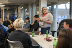 A woman standing holding a book, presenting to a group of people sitting at tables and on couches.
