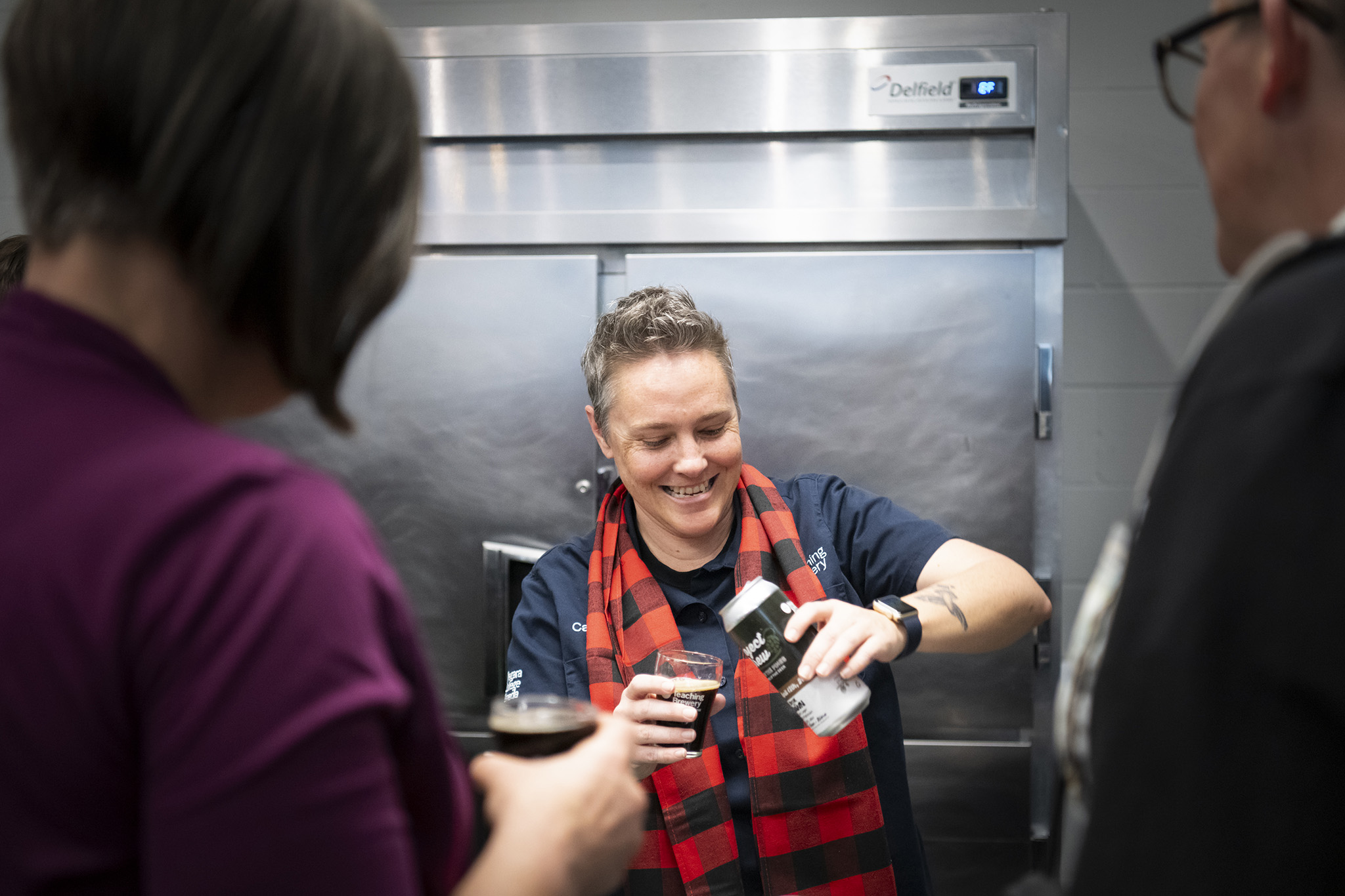 Two people watching a person pour a can of beer into a glass.