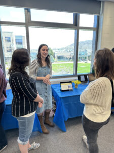 Three women engaging in conversation, standing in front of a large window and a table with a blue tablecloth with laptops and florals on it.