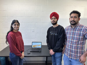 One woman (left) and two men (right) standing around a table with a laptop on it.