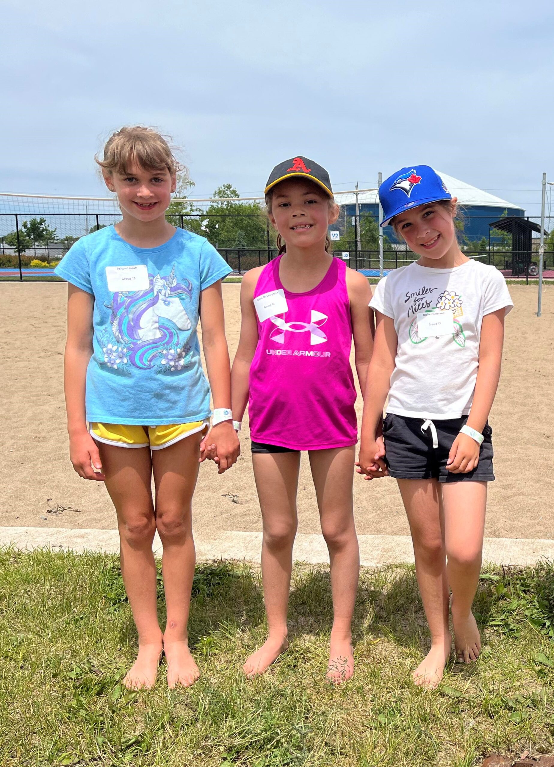 three young girls pose for a photo on the edge of a beach volleyball court