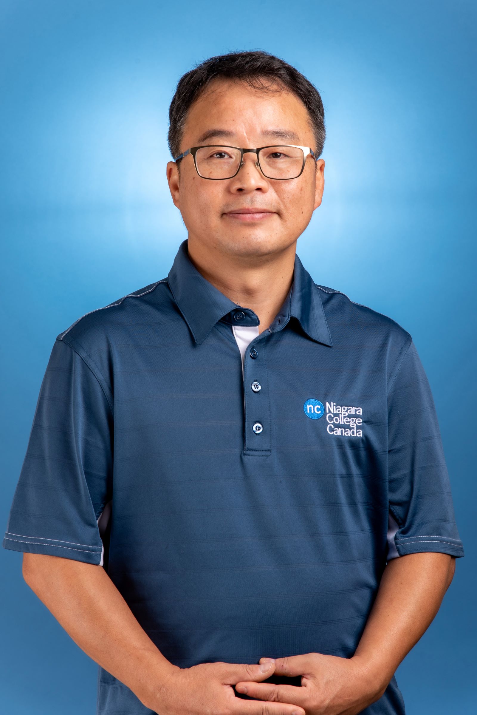 a head shot of a man with short dark hair and glasses wearing a blue NC polo t-shirt