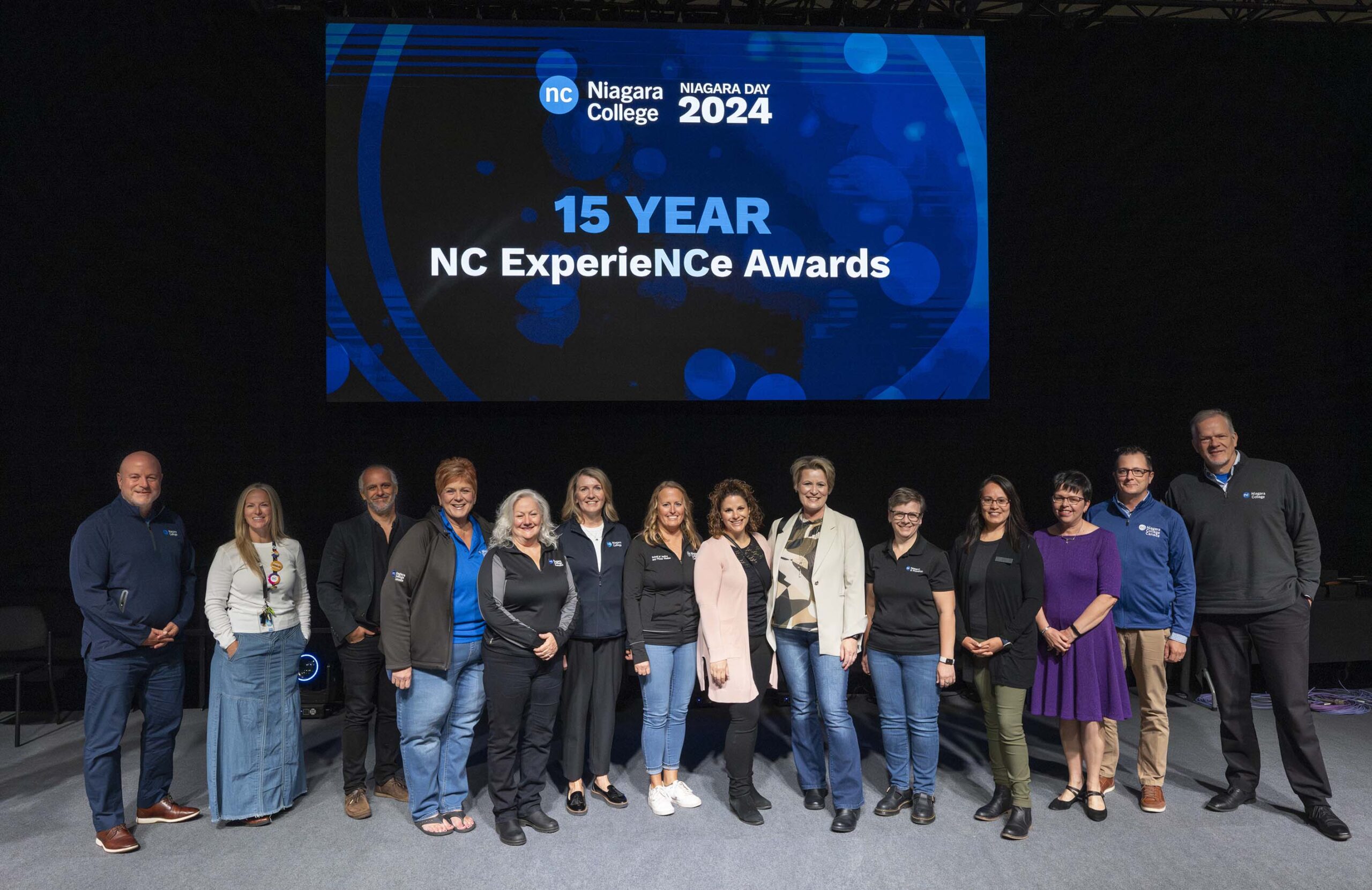 a group of people stand on stage below a digital screen
