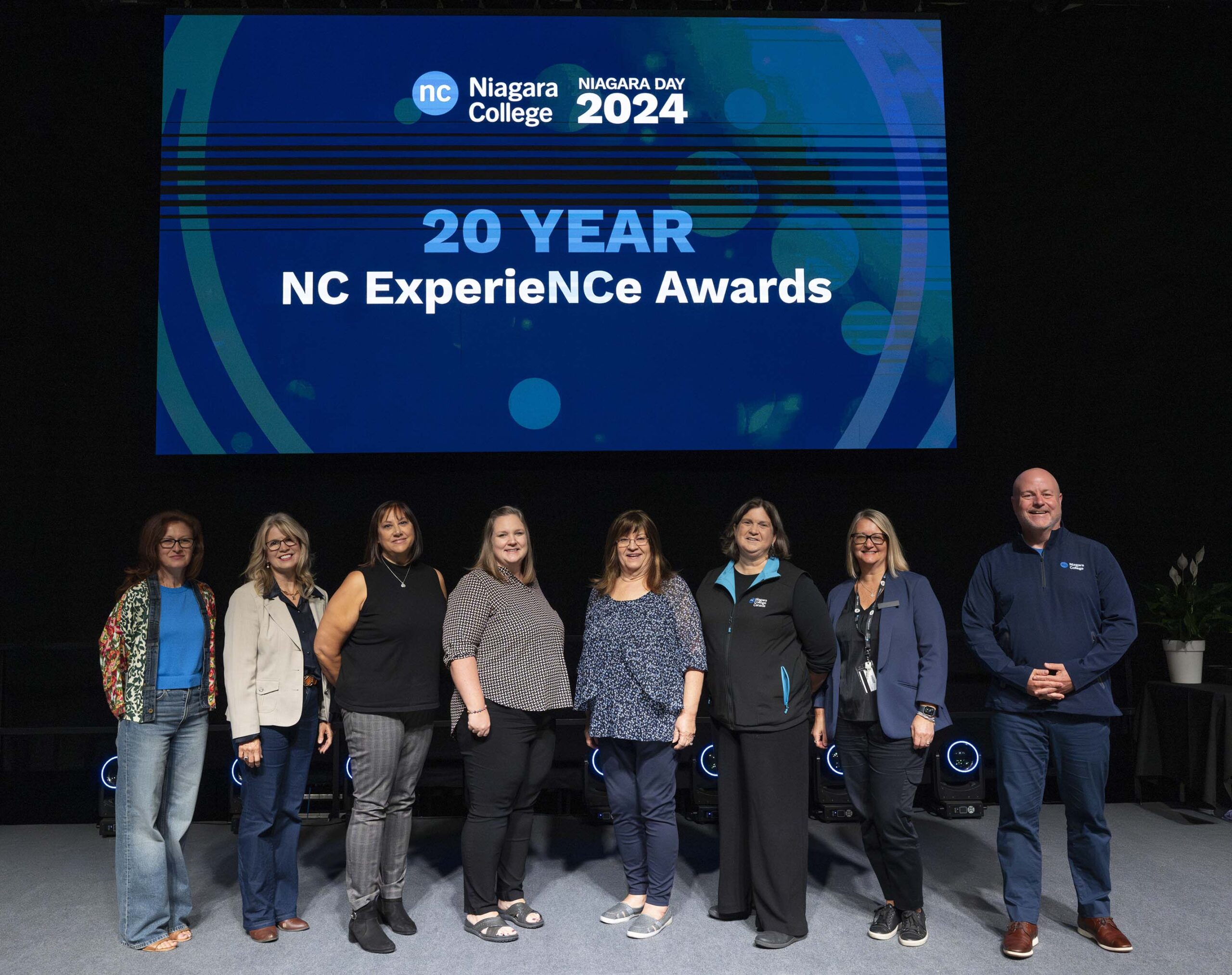 a group of people stand on stage below a digital screen