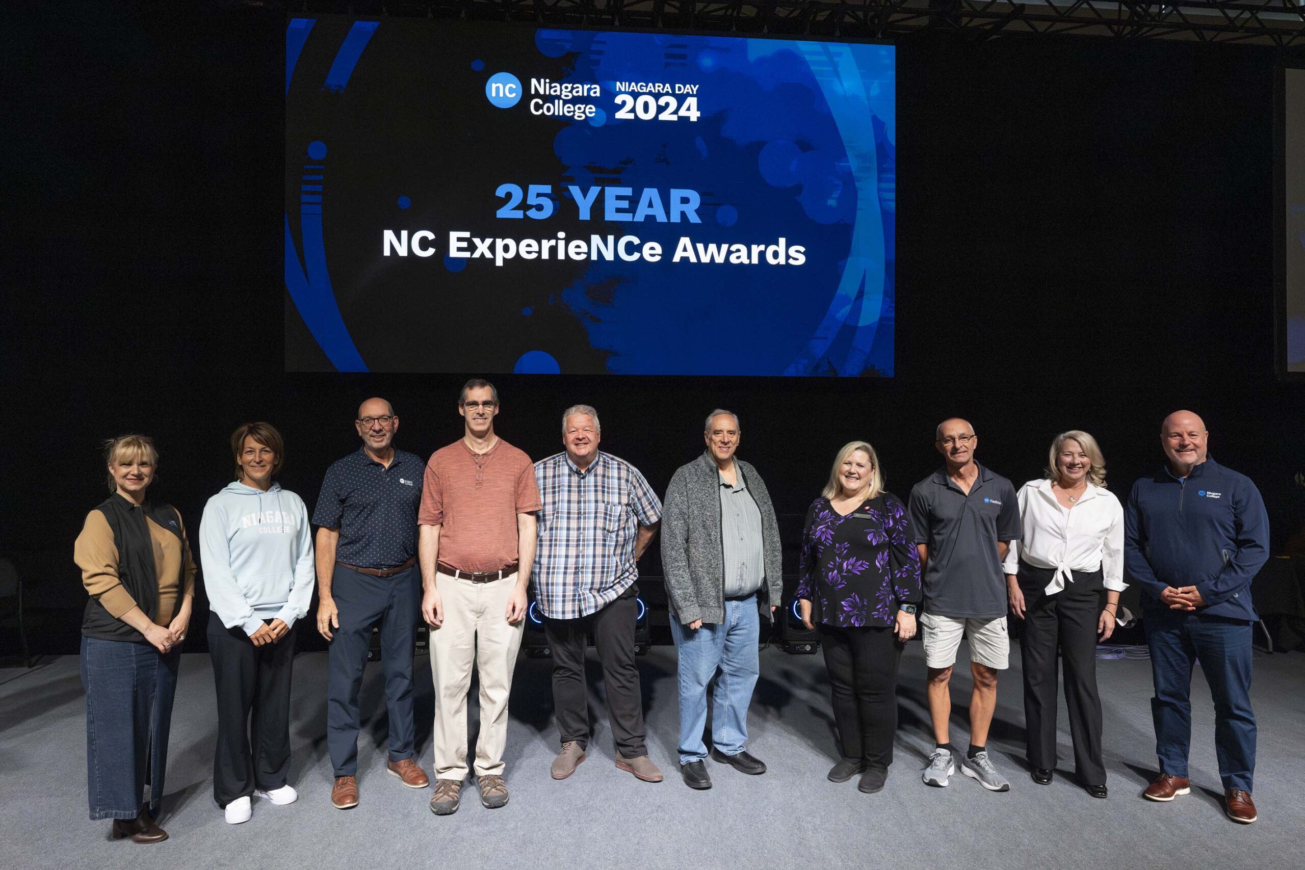 a group of people stand on stage below a digital screen