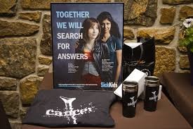 A table with two mugs, a t-shirt that reads Cancer, and a poster that reads together we will search for answers, with two girls in the background.