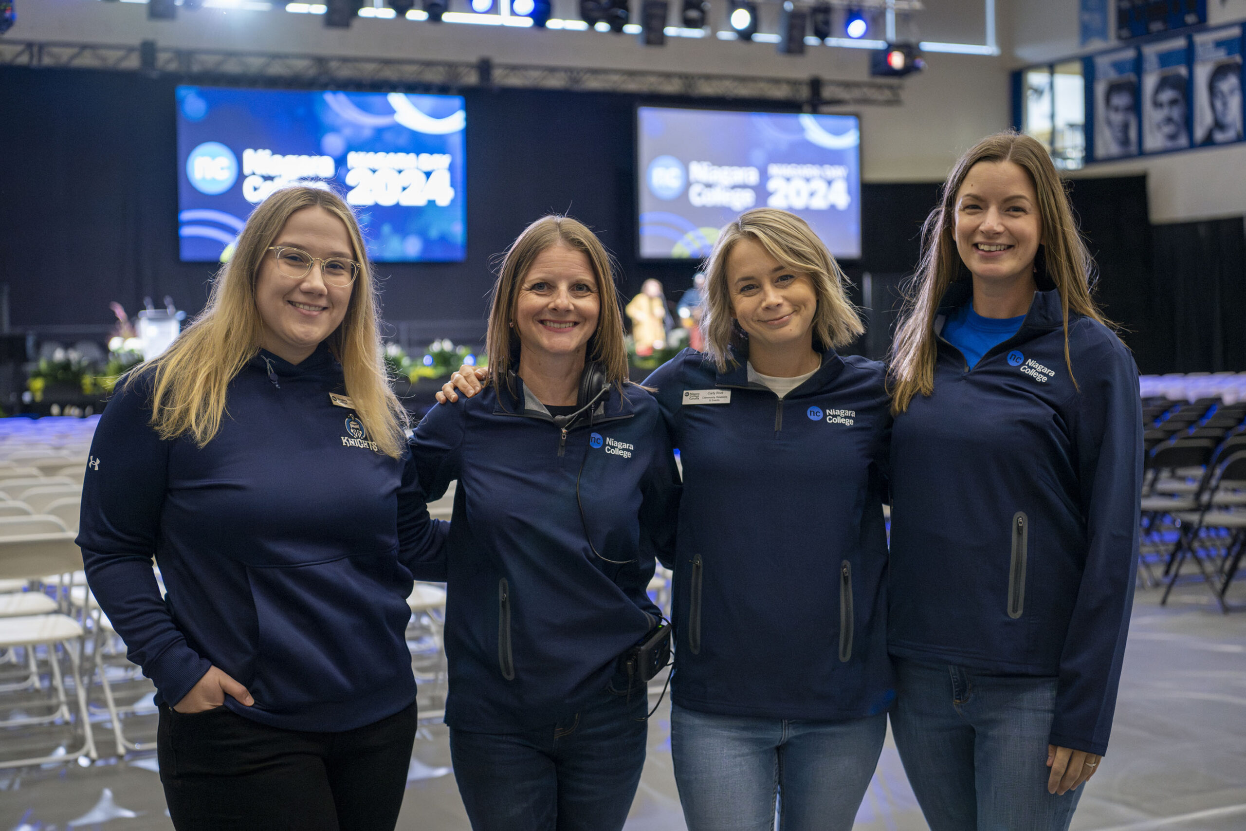 Four women wearing branded blue college sweaters stand side by side with their arms around each other
