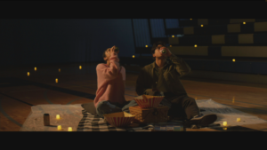 A screenshot of a scene in a film where a young man and woman sit on a blanket inside a gymnasium, having a picnic while drinking from a can