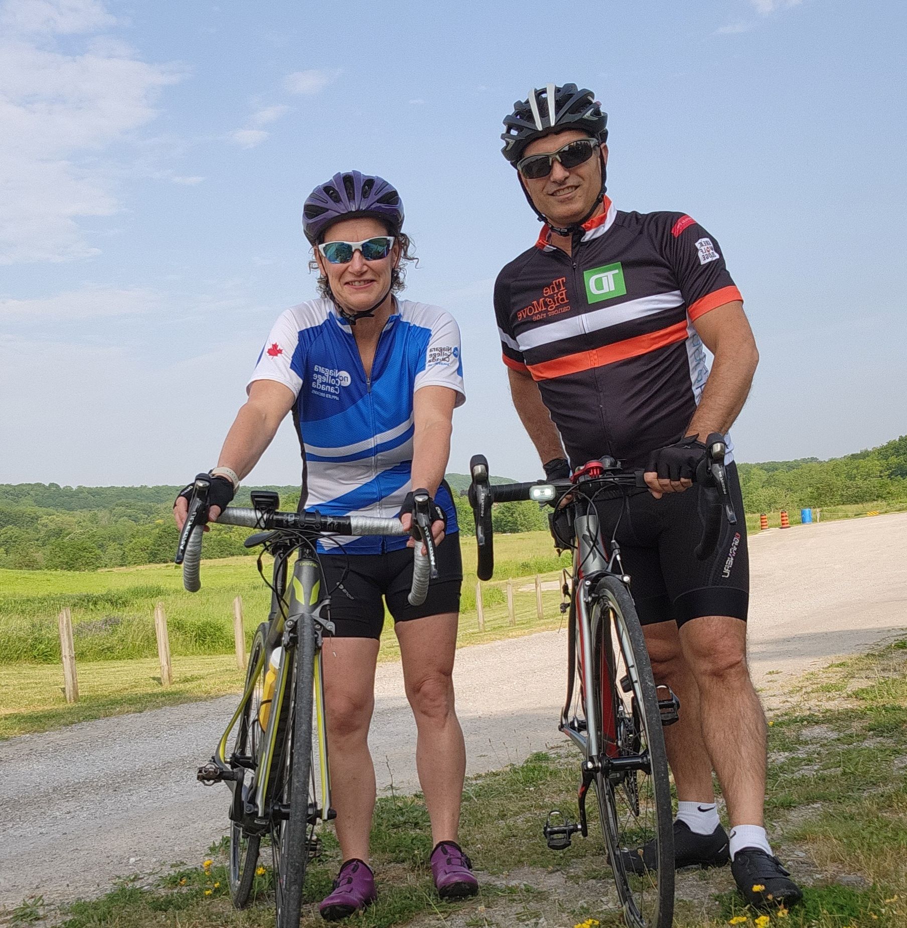 two cyclists wearing bike gear and helmets stand beside their bikes on the side of a country road