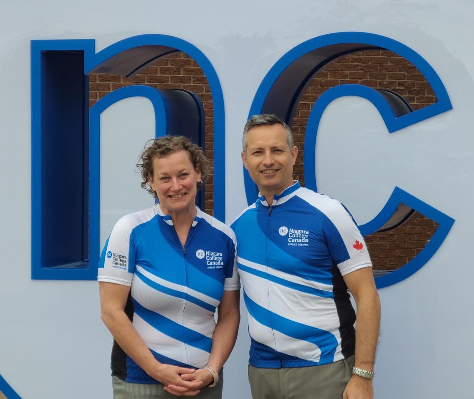 two people wearing blue and white Niagara College cycling jerseys pose for a photo in front a a large blue and white NC sign