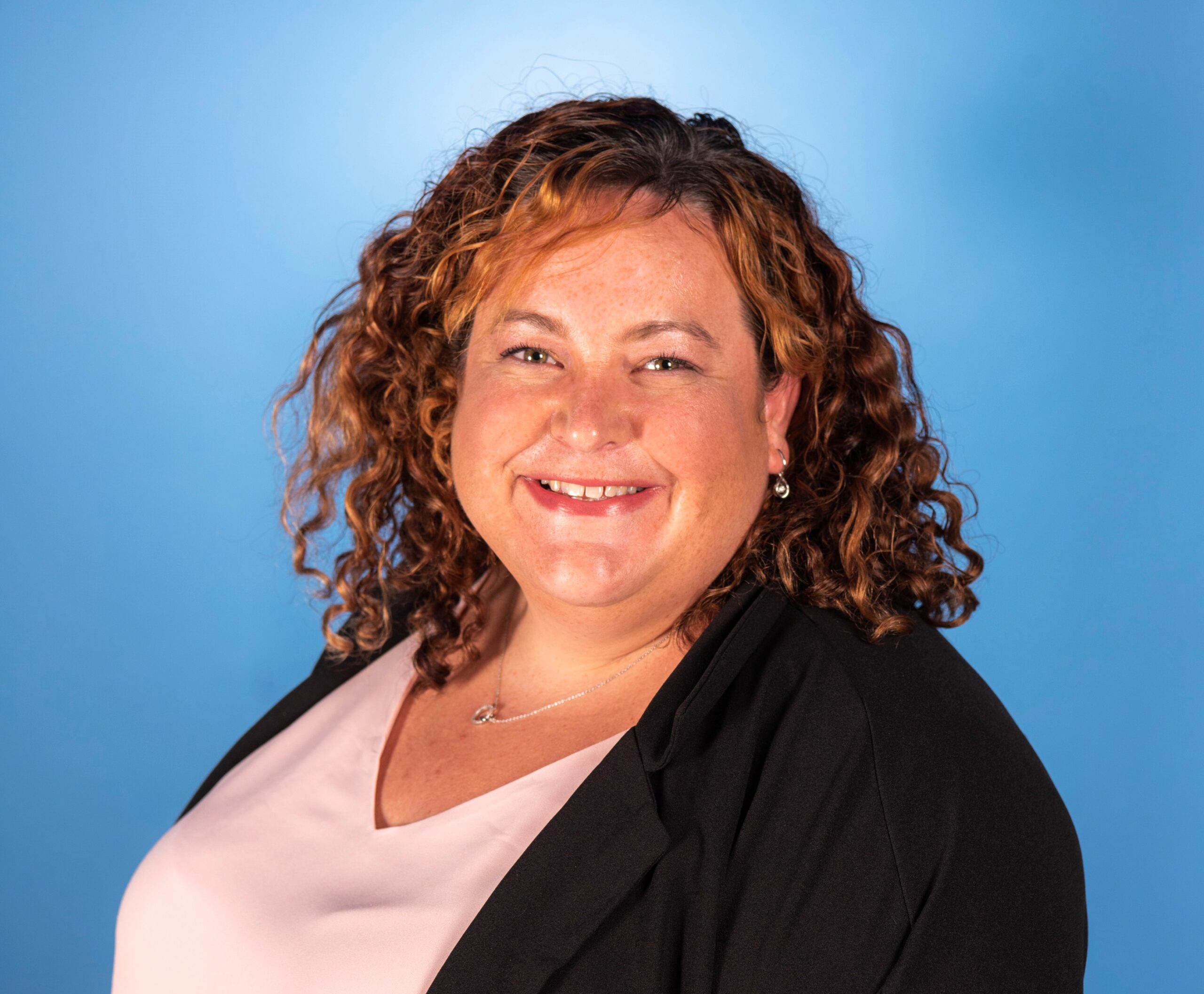 A woman with curly brown hair is smiling in front of a blue background. She is wearing a pink and black shirt.