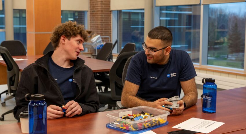 two students sit at a table talking with a container of lego in front of them