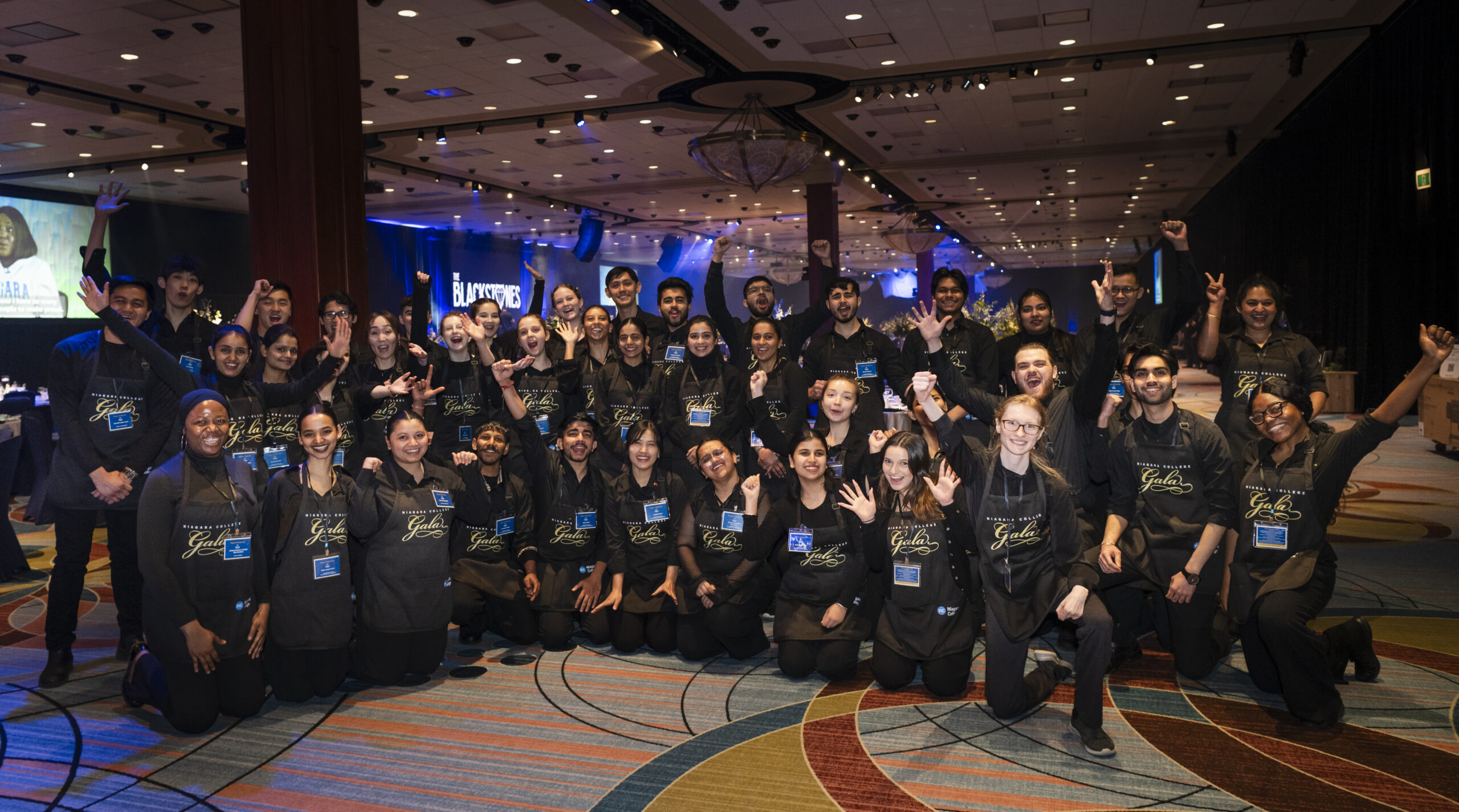 A large group of gala volunteer students in matching black outfits and aprons are smiling and waving in the venue hall.