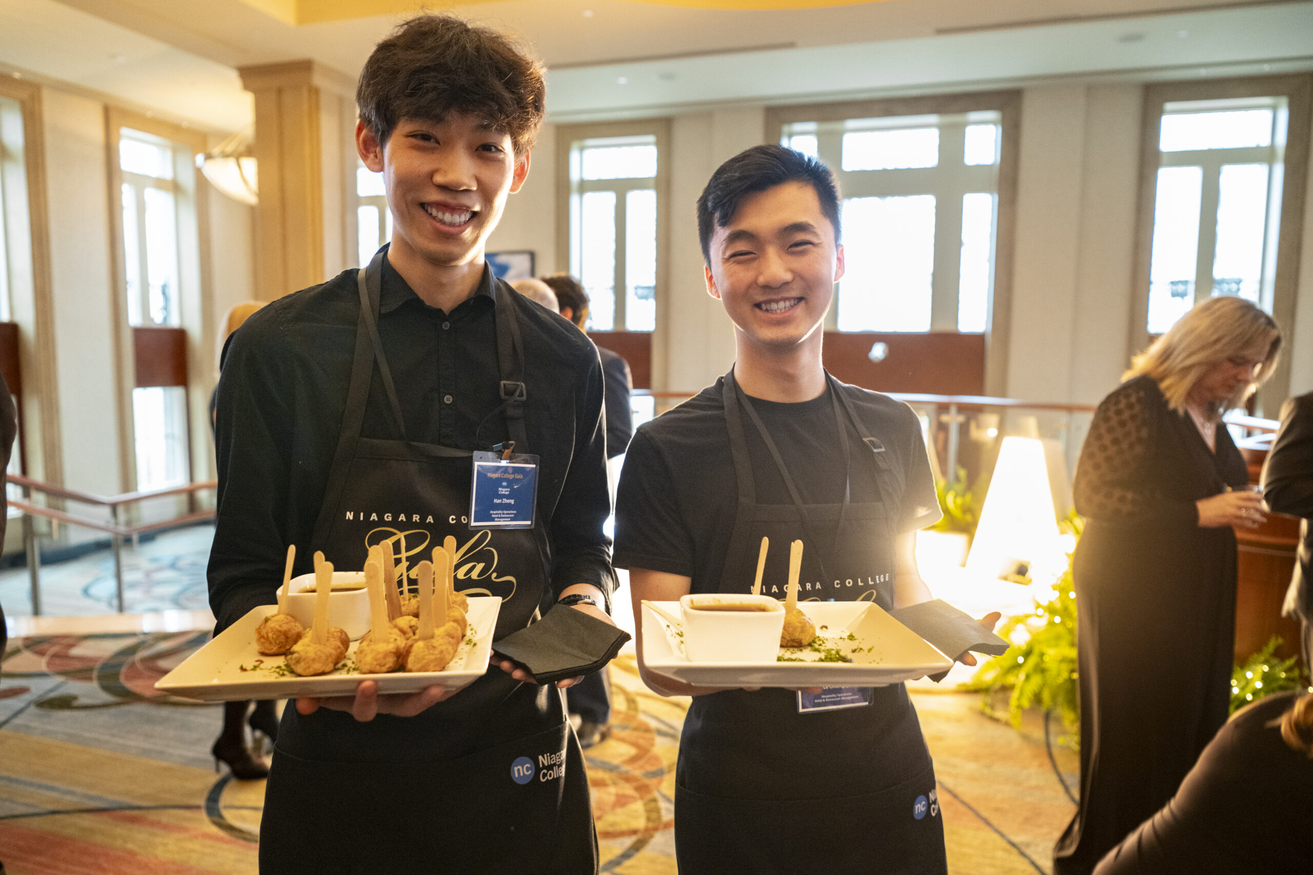 Two young male students in black aprons smiling, holding trays of gourmet appetizers.