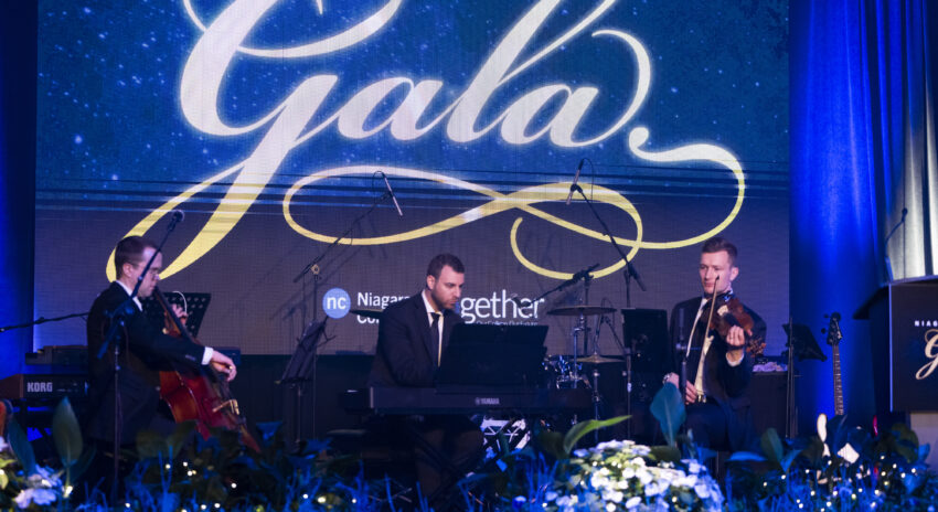 A musical trio performs on stage, featuring a pianist, cellist, and violinist in formal attire against a backdrop with the word "Gala."