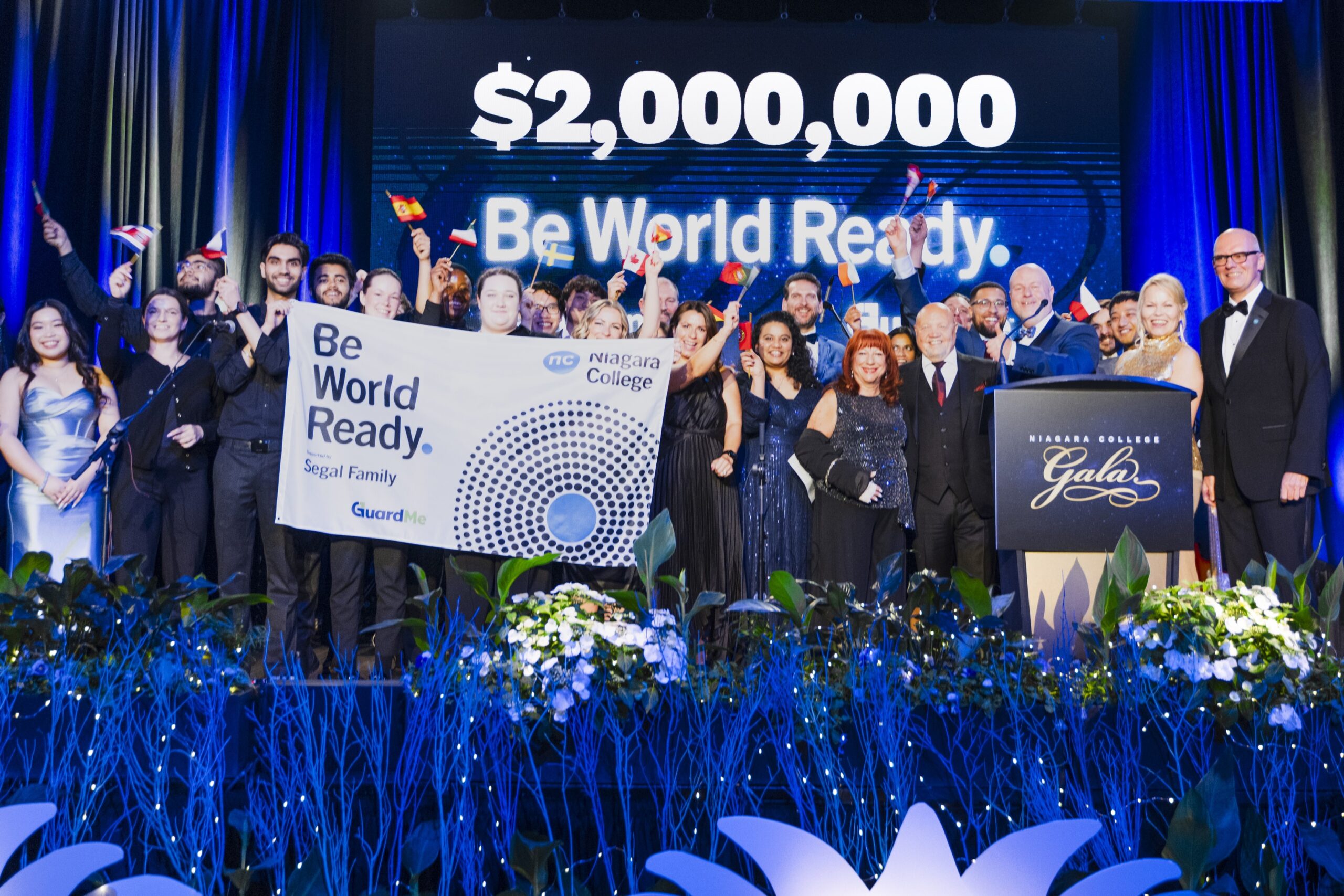 Group of people in formal attire on stage at a gala, celebrating a $2,000,000 donation. They hold flags and a "Be World Ready" banner under bright blue lighting.