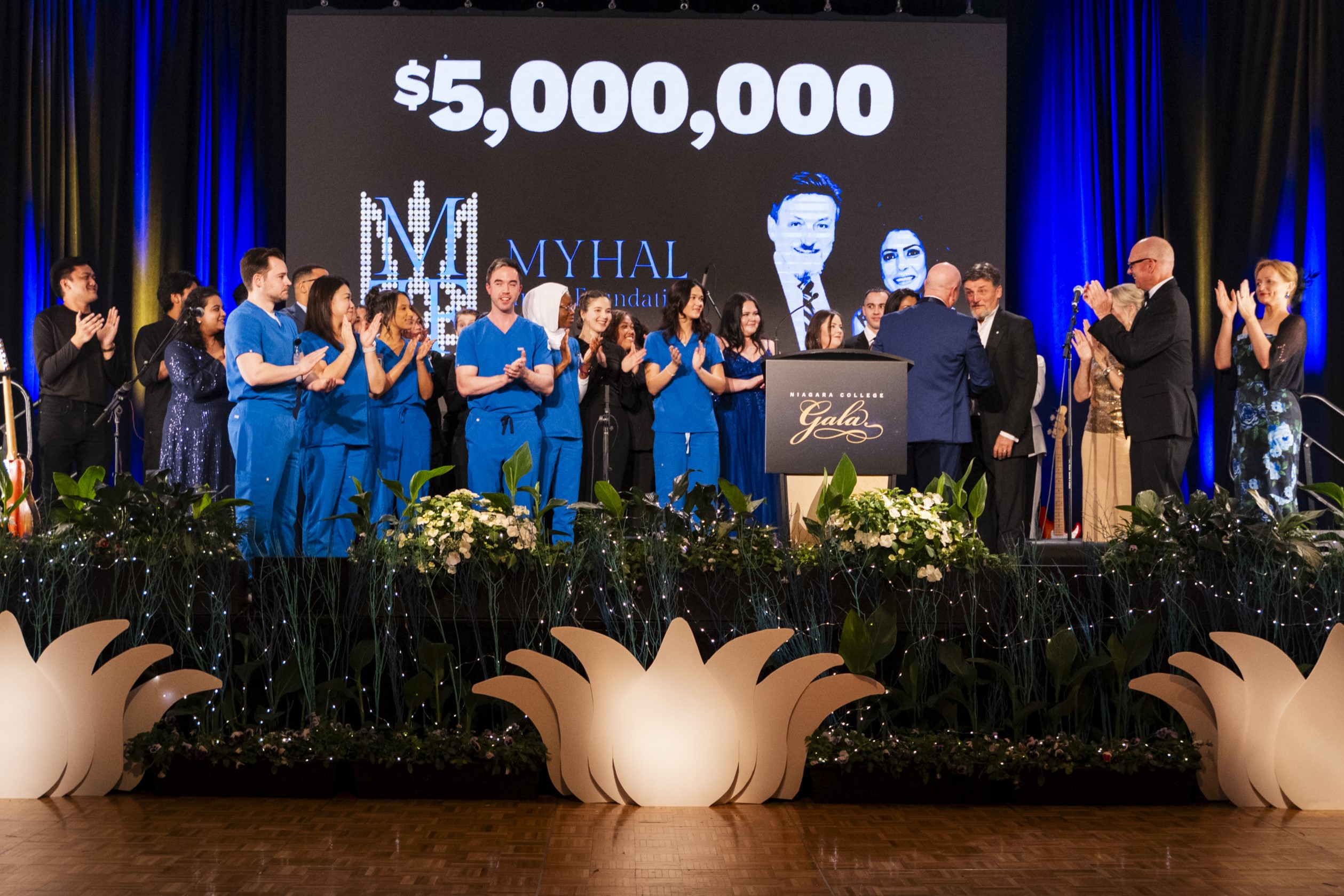 A group celebrates on stage at a gala, applauding the announcement of a $5 million donation, as medical students in blue scrubs stand near a podium with flowers.