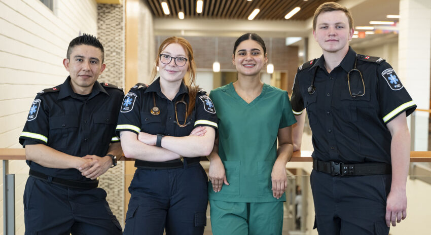 three students wearing paramedic uniforms and one student wearing green scrubs lean against a railing inside a campus building