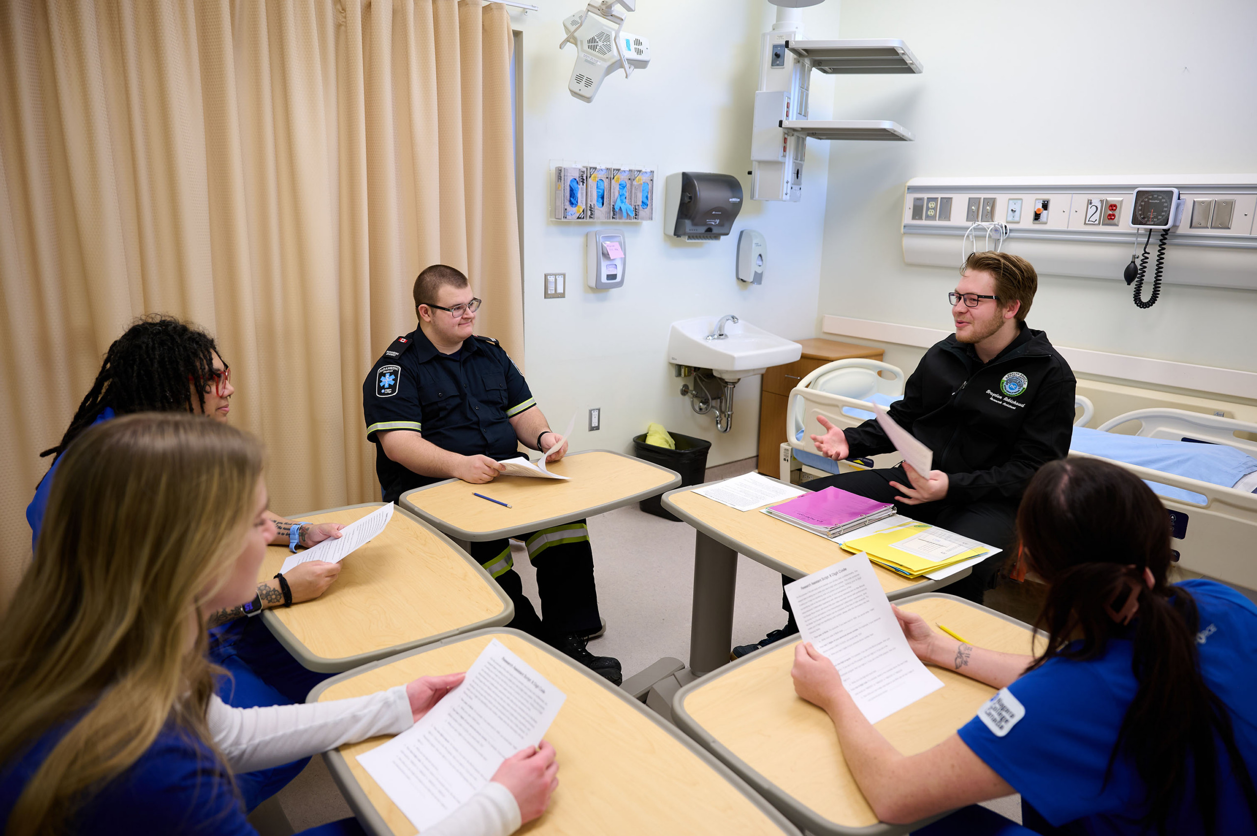five students sit at small tables grouped together in a patient room