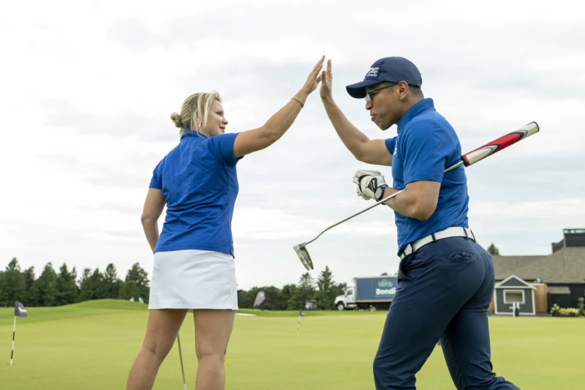 A woman in a golf outfit and a man in a blue golf shirt exchange a high-five on a putting green.