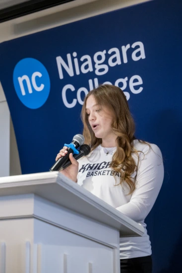 A female student-athlete wearing a white long-sleeve Niagara Knights Basketball shirt speaks at a wooden podium, holding a microphone in front of a blue Niagara College backdrop.