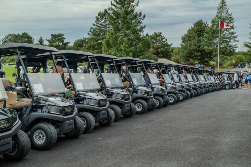A long row of black golf carts lined up on a paved path at a golf course with a Canadian flag waving in the background.