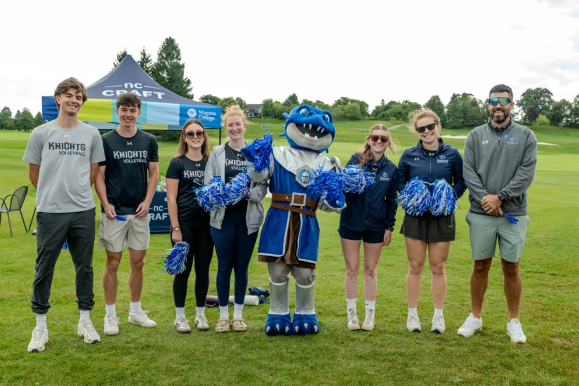 A group of Niagara College Knights athletes and their mascot pose together on a golf course.