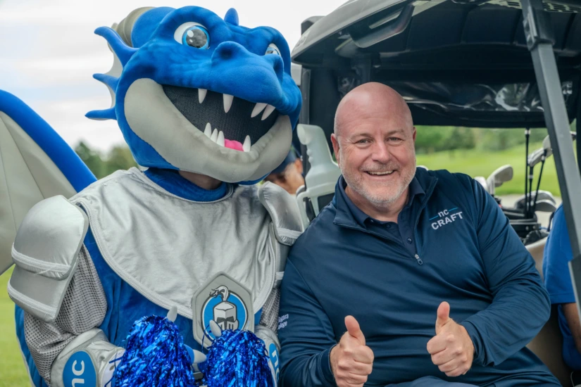 NC President Sean Kennedy and Knights mascot Noble give a thumbs-up while sitting side by side in a golf cart.